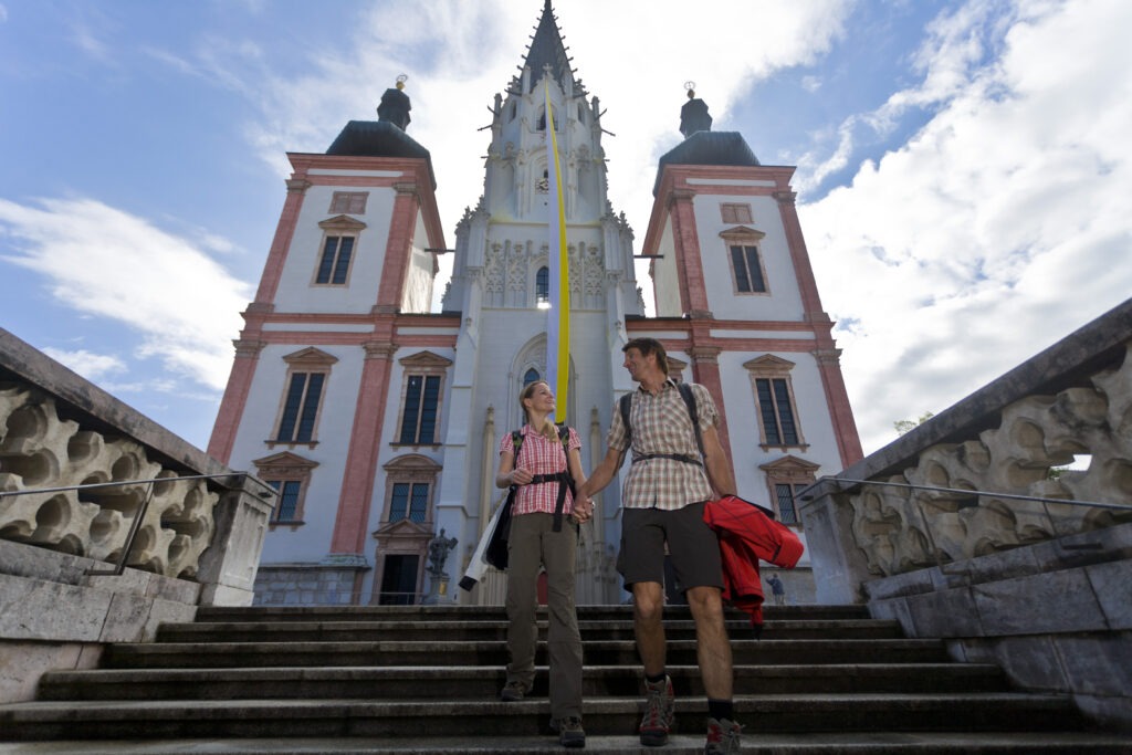 Zwei Wandernde stehen auf den Stufen vor der Basilika Mariazell und blicken einander an. Hinter ihnen erhebt sich die große Kirchenfassade mit ihren zwei Türmen und dem hohen gotischen Mittelturm vor einem hellen Himmel mit Wolken.