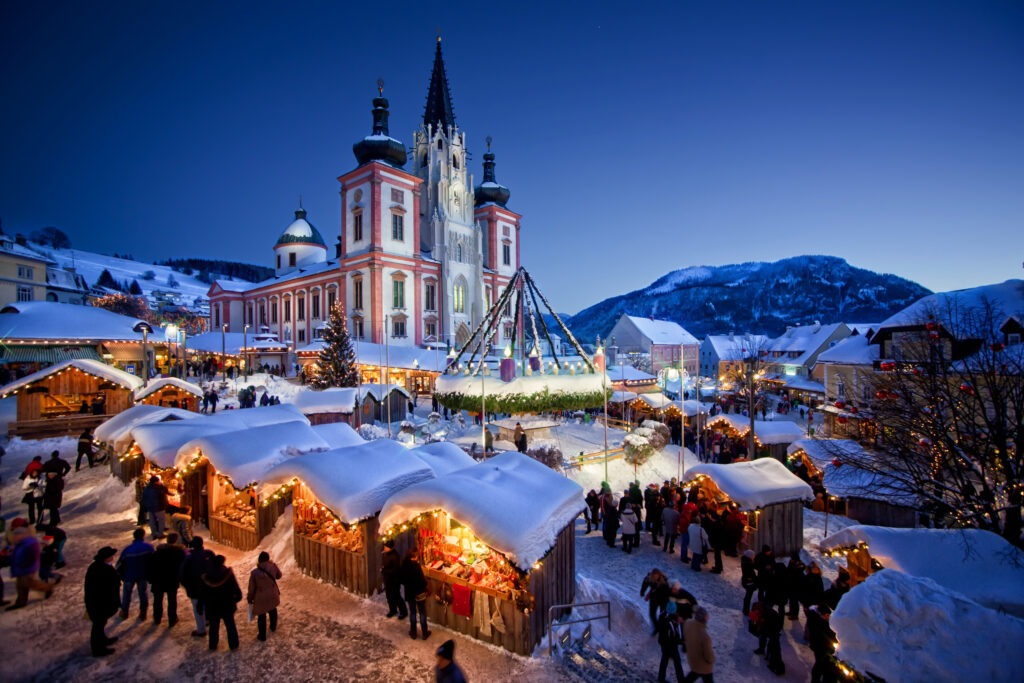 Verschneiter Mariazeller Advent in der blauen Stunde mit beleuchteten Adventhütten, großem Adventkranz und der Basilika Mariazell im Hintergrund.